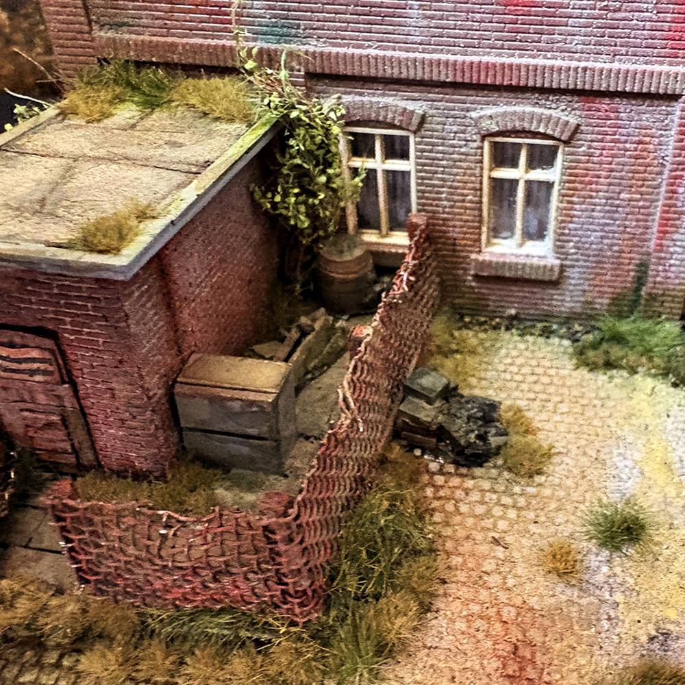 Verwinkelter Hinterhof mit Backsteingebäuden, Schuppen und überwuchertem Maschendrahtzaun